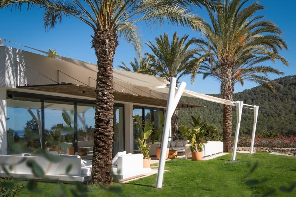 This luxurious modern house features large windows and a sleek white exterior, complemented by a shaded patio. Surrounded by tall palm trees and lush green grass, it offers breathtaking views of the hills, perfect for luxury travel seekers under the clear blue sky. David Pearson Travel
