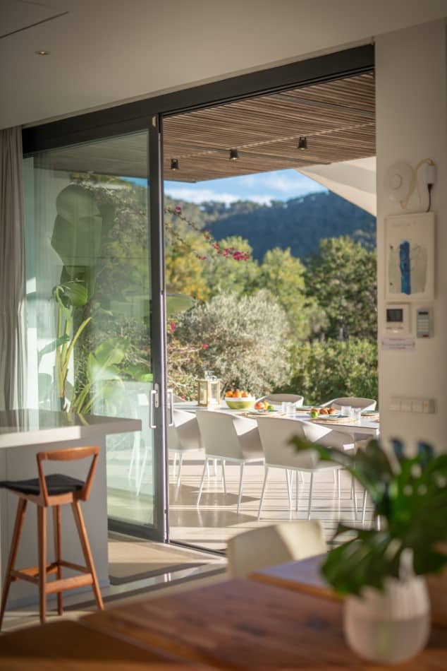 A modern dining area with a wooden table and chairs leads to a sunlit patio through a glass sliding door. The patio, reminiscent of luxury travel destinations, boasts a large dining table set with dishes, surrounded by nature with views of trees and distant hills. David Pearson Travel