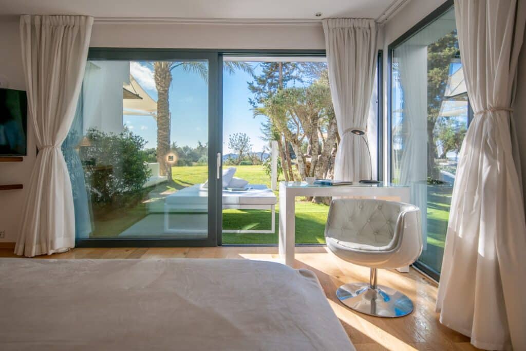 A bright, modern bedroom with floor-to-ceiling glass doors opens onto a garden view, ideal for luxury travel experiences. The room features white curtains, a sleek white chair, and a small desk. Outside, greenery and a hint of sunny sky create a serene ambiance. David Pearson Travel