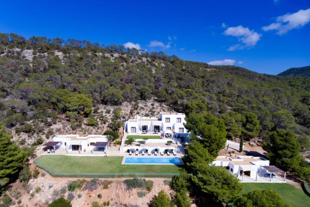 Aerial view of a luxury travel destination: a large white villa nestled among lush green trees and hills. The villa boasts a swimming pool, sun loungers, and a manicured lawn under the clear blue sky. Multiple white buildings enhance the opulent allure of the property. David Pearson Travel