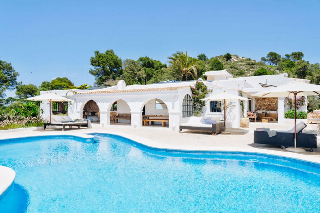 A Mediterranean-style villa, perfect for luxury travel, boasts white arches and terracotta roof tiles nestled amid lush greenery. It features a lavish outdoor pool with sun loungers and patio umbrellas under the clear blue sky, offering an idyllic escape. David Pearson Travel