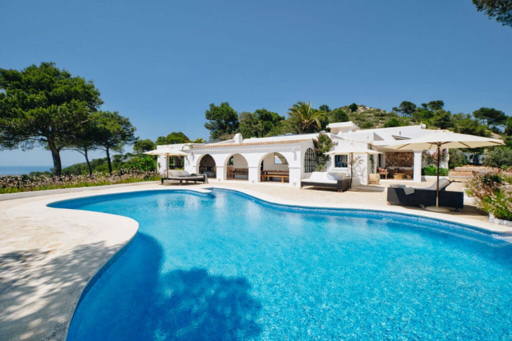 Luxury villa with white arches and stone accents, a dream destination for luxury travel enthusiasts. Surrounded by trees, a sparkling blue swimming pool reflects the clear sky. Outdoor lounge seating and umbrellas elegantly arranged around the pool enhance your lavish escape. David Pearson Travel