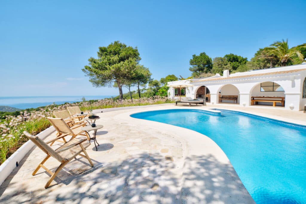 A scenic view of a luxurious patio with a curved pool and lounge chairs. The patio overlooks a vast ocean, surrounded by lush trees and flowering plants. A white villa with arches and outdoor seating peeks through under the clear blue sky, epitomizing luxury travel at its finest. David Pearson Travel