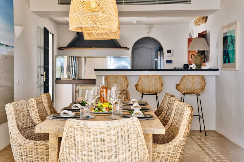 A touch of luxury travel ambiance graces the dining room, featuring a wooden table set for six, embraced by wicker chairs. A bottle and a dish with greens adorn the table. Behind, the kitchen boasts a counter with rattan bar stools, illuminated by woven pendant lights hanging elegantly from above. David Pearson Travel