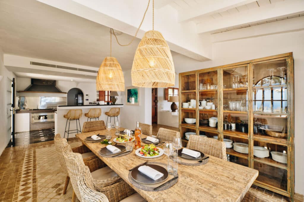 A dining room with a rustic wooden table set for a meal, surrounded by wicker chairs. Two woven pendant lights hang above, exuding an air of luxury travel charm. In the background, theres a glass cabinet with dishes and a kitchen area featuring bar stools and modern appliances. David Pearson Travel