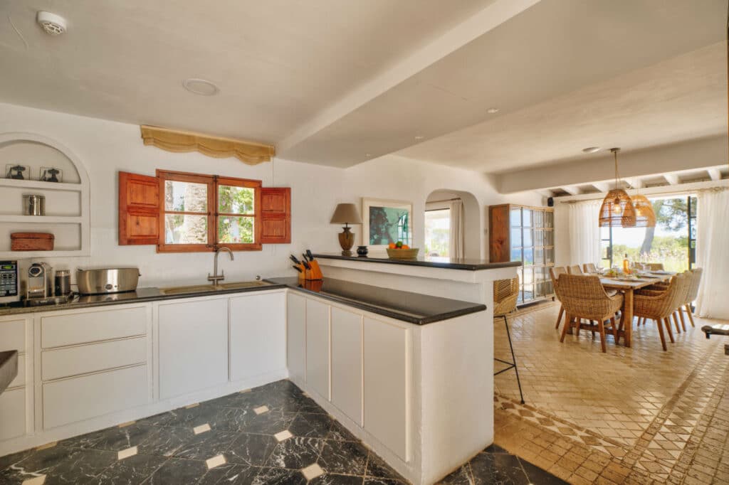 A bright kitchen with white cabinets, black countertops, and red shutters over the window opens to a dining area featuring a long table with wicker chairs and a luxury pendant light. Large windows provide a travel-inspired view of the trees outside. David Pearson Travel
