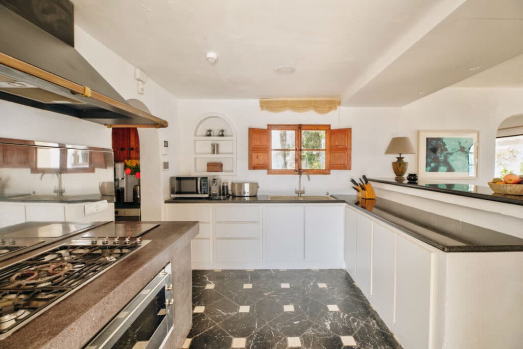 A bright kitchen with white cabinetry, a black and white tiled floor, and wooden window shutters evokes a luxury travel vibe. The room features a gas stovetop and oven on a central island, an inviting sink under the window, state-of-the-art appliances, and an elegant fruit bowl on the counter. David Pearson Travel