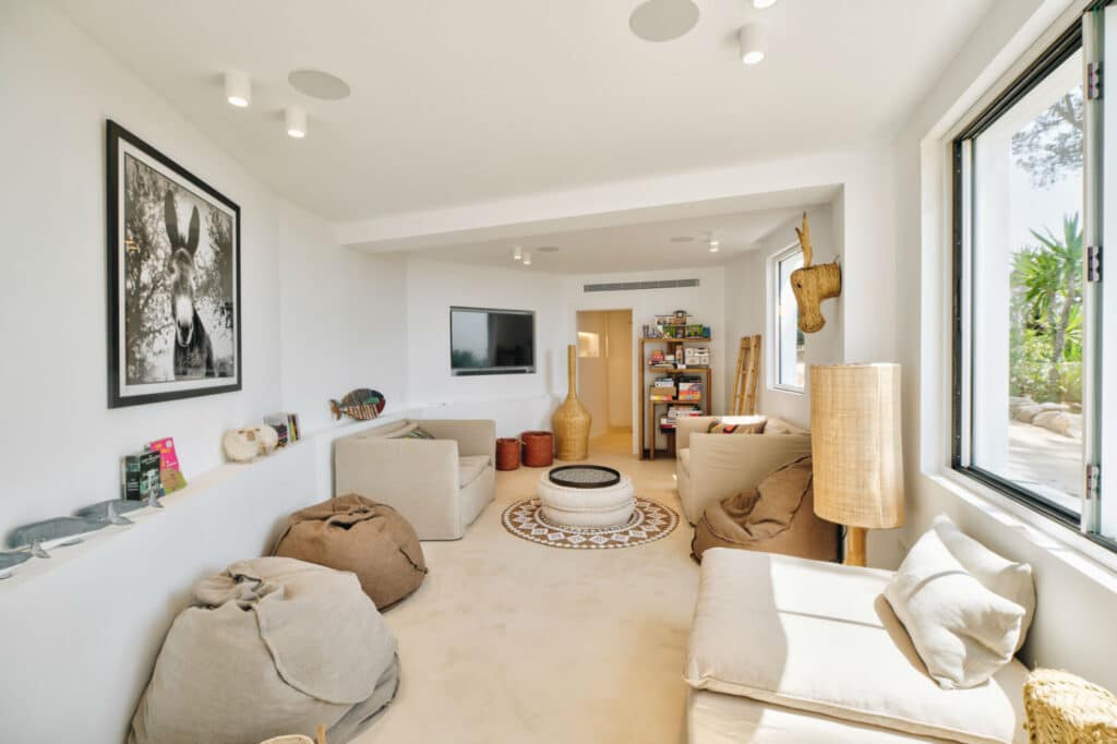 A cozy living room with beige furniture, including armchairs and a bean bag that evoke the comfort of luxury travel. A large window lets in natural light. Walls are adorned with art, and a TV is mounted on the wall. A woven rug and various decorative items add warmth to the space. David Pearson Travel