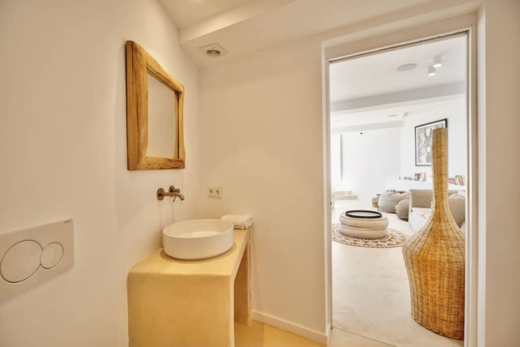 A small, bright bathroom with a round vessel sink and wooden-framed mirror exudes luxury. A wicker vase graces the floor by the entrance, seamlessly leading to a cozy living room featuring a large beige sofa with natural light streaming in—a perfect retreat after indulgent travel adventures. David Pearson Travel