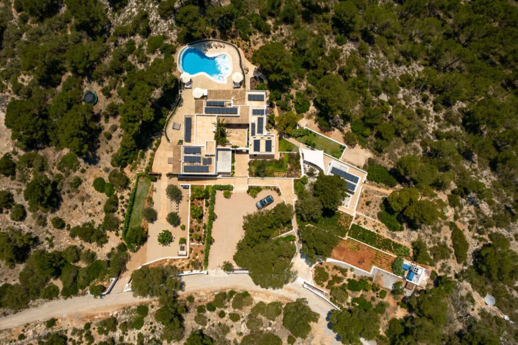 Aerial view of a large, modern villa perfect for luxury travel, surrounded by dense trees. The property boasts a swimming pool, flat-roofed buildings, a central courtyard, and a car parked on the driveway. The surrounding landscape is beautifully rocky and green. David Pearson Travel