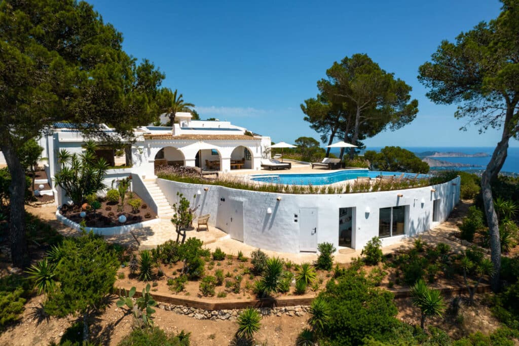 An aerial view reveals a luxurious Mediterranean villa with an arched terrace and pool, nestled among pine trees. This oasis overlooks a scenic coastline and blue sea under a clear sky, perfect for luxury travel enthusiasts. The landscape is enriched with greenery and charming stone paths. David Pearson Travel