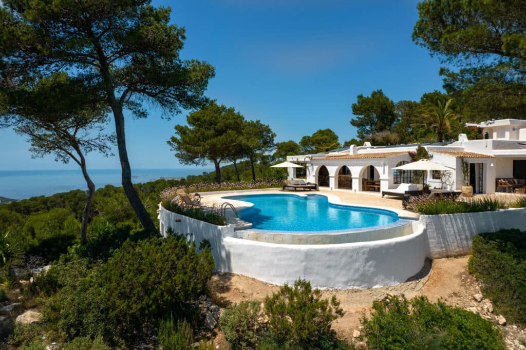 A luxurious white villa, perfect for luxury travel, features arched windows overlooking an expansive swimming pool surrounded by lush greenery and pine trees. In the background, theres a clear blue sky and an ocean view. David Pearson Travel