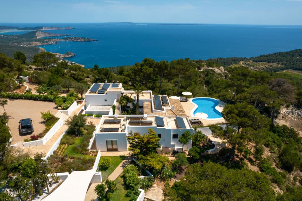 Aerial view of a modern white villa epitomizing luxury travel, with a uniquely shaped pool. Surrounded by lush greenery and overlooking a scenic coastline with azure seas and distant islands under a clear sky, the property also features a driveway with a sleek car parked. David Pearson Travel