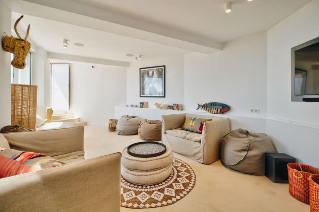 A cozy living room with beige sofas and poufs, adorned with colorful cushions, offers a touch of luxury travel charm. A round, patterned rug centers the space. Wall art includes a fish sculpture and framed picture. Natural light fills the room, enhancing the elegance of a mounted flat-screen TV. David Pearson Travel