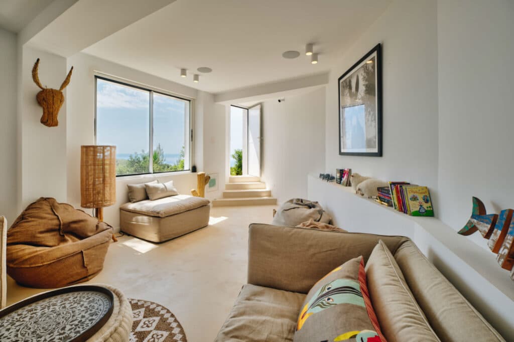 Sunlit living room with beige sofas and a large window. Wall shelves hold books and decor. A wicker animal head decorates the wall, adding a touch of luxury travel charm. A rug and pillows add coziness. Stairs lead to a slightly raised area by the window, showing a glimpse of greenery outside. David Pearson Travel