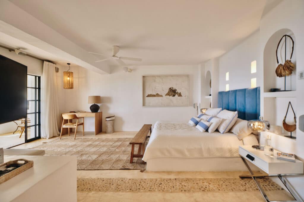 Spacious, well-lit bedroom with a luxury travel vibe, featuring a large bed adorned with blue pillows, a wooden bench at the foot, and modern decor. A work desk by the window and a TV on the left complete this serene retreats neutral tones and open design. David Pearson Travel