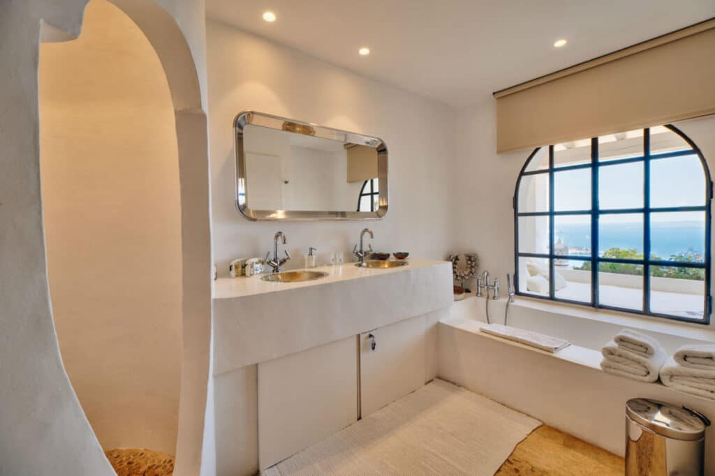 A minimalist bathroom with a curved, white structure and a large window overlooking the ocean embodies luxury travel. It features a double sink with a modern mirror above, a bathtub adorned with neatly folded towels, and natural stone flooring. David Pearson Travel