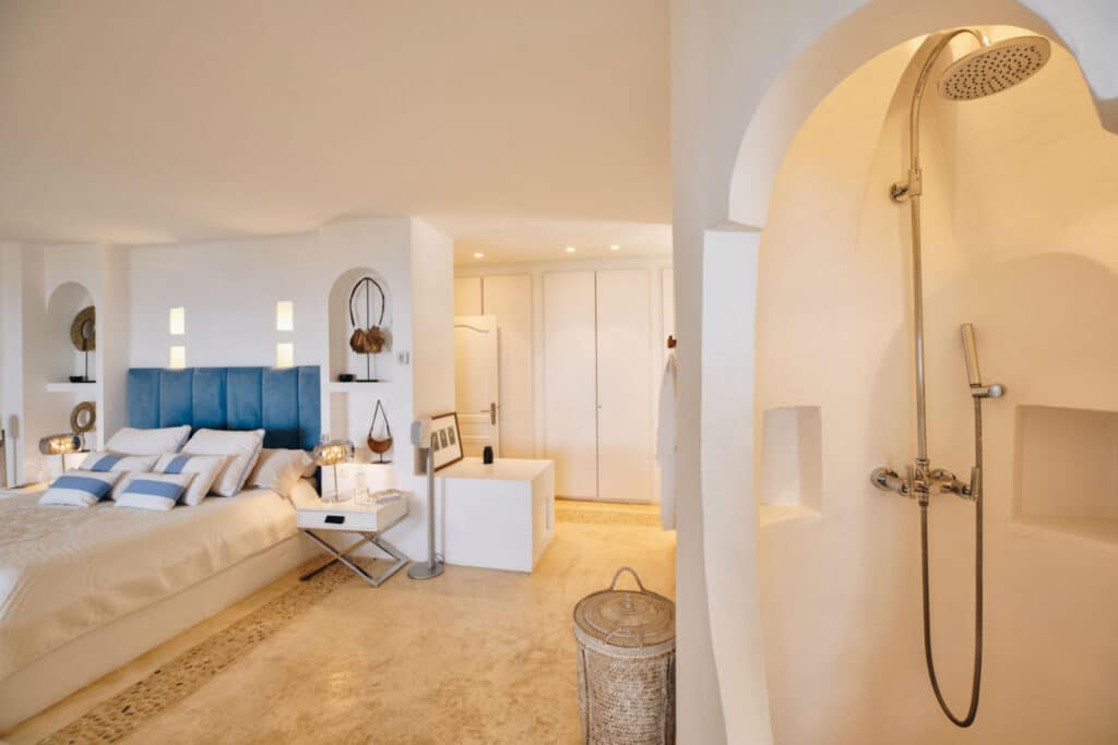 A spacious, elegantly decorated bedroom perfect for luxury travel, featuring a large bed with a blue headboard and crisp white bedding. The rooms warm lighting complements its stone-tiled floor, archway shower area, and chic wicker laundry basket. David Pearson Travel