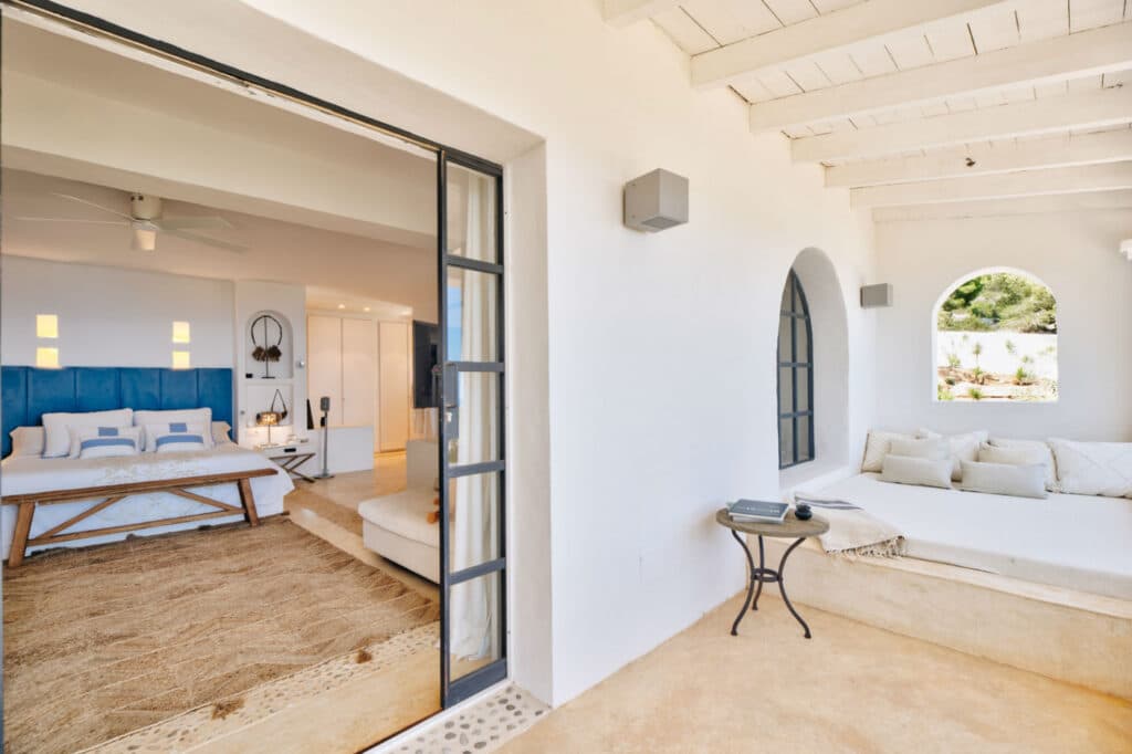 A bright, airy bedroom, ideal for luxury travel enthusiasts, opens to a cozy outdoor seating area. The room features a large bed with blue accents, natural woven rugs, and white walls. Outside, a built-in bench with cushions and arched windows enhance the serene ambiance. David Pearson Travel