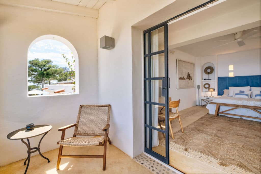 Cozy room with open sliding doors to a small patio, perfect for luxury travel enthusiasts. A woven chair and round table sit on the patio. Inside, a rug, wooden chair, and a bed with a blue headboard complement the wall decor. An arched window provides a serene view of the trees outside. David Pearson Travel