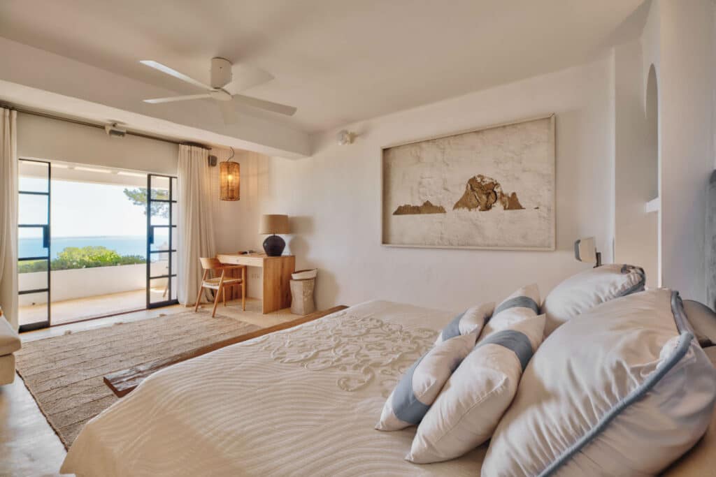 Bright and airy bedroom with a large bed adorned with white and blue pillows, perfect for luxury travel indulgence. A desk and chair sit near glass doors opening to a balcony offering a sea view. A ceiling fan and artwork enhance the serene, minimalist decor. David Pearson Travel
