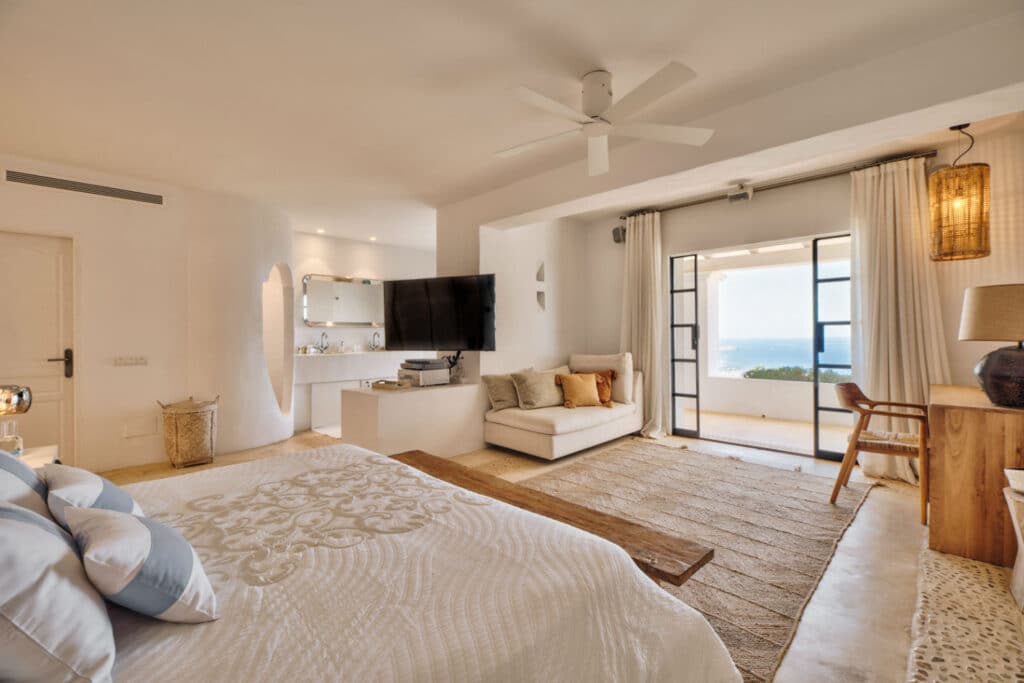 Cozy bedroom with white walls and ceiling fan, perfect for luxury travel. Features a large bed with patterned bedding, wooden furniture, and a TV on a cabinet. A window with curtains reveals an ocean view. Neutral tones and soft lighting create a relaxing atmosphere. David Pearson Travel