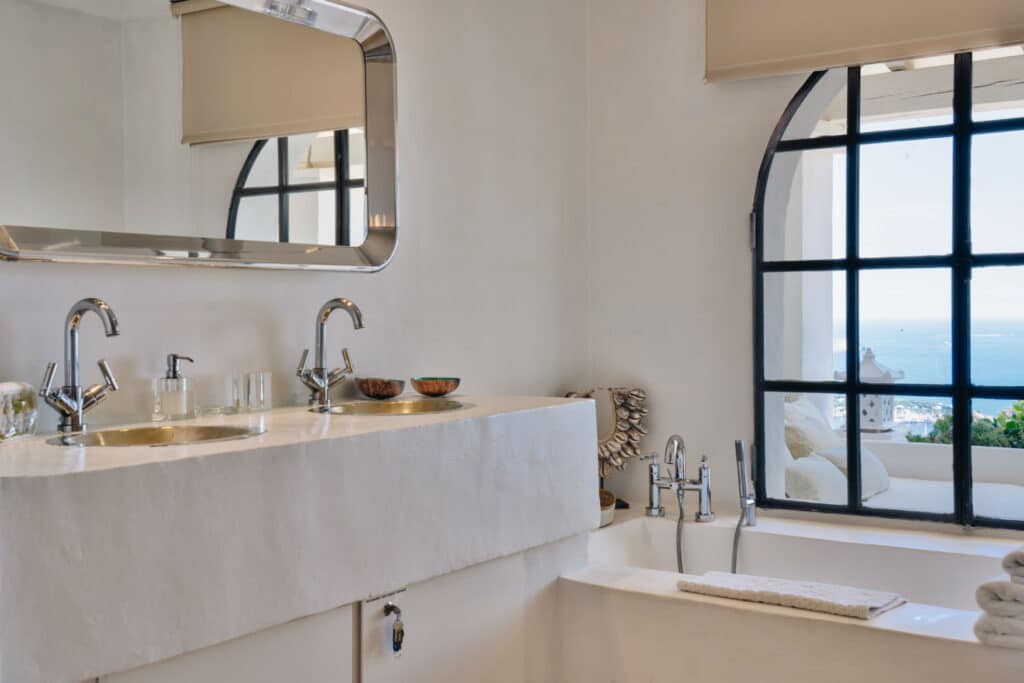 A modern bathroom exuding luxury travel vibes features a large white sink with dual faucets and a mirror. Nearby, a bathtub awaits, accompanied by a folded towel. A grand arched window offers views of the sea and outdoor seating, while neutral tones enhance the serene ambiance. David Pearson Travel