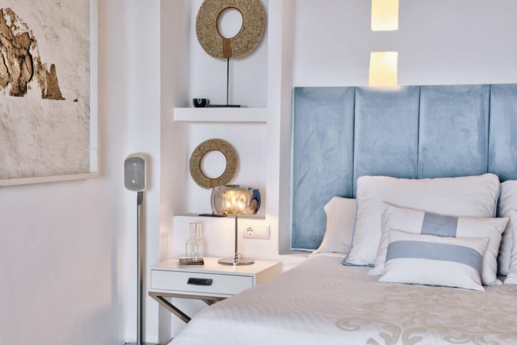 A modern bedroom with a cushioned blue headboard, white bedding, and multiple pillows evokes the elegance of luxury travel. A white nightstand holds a metallic lamp, while decorative round mirror accents adorn the wall. A floor lamp and framed abstract art complete the decor. David Pearson Travel