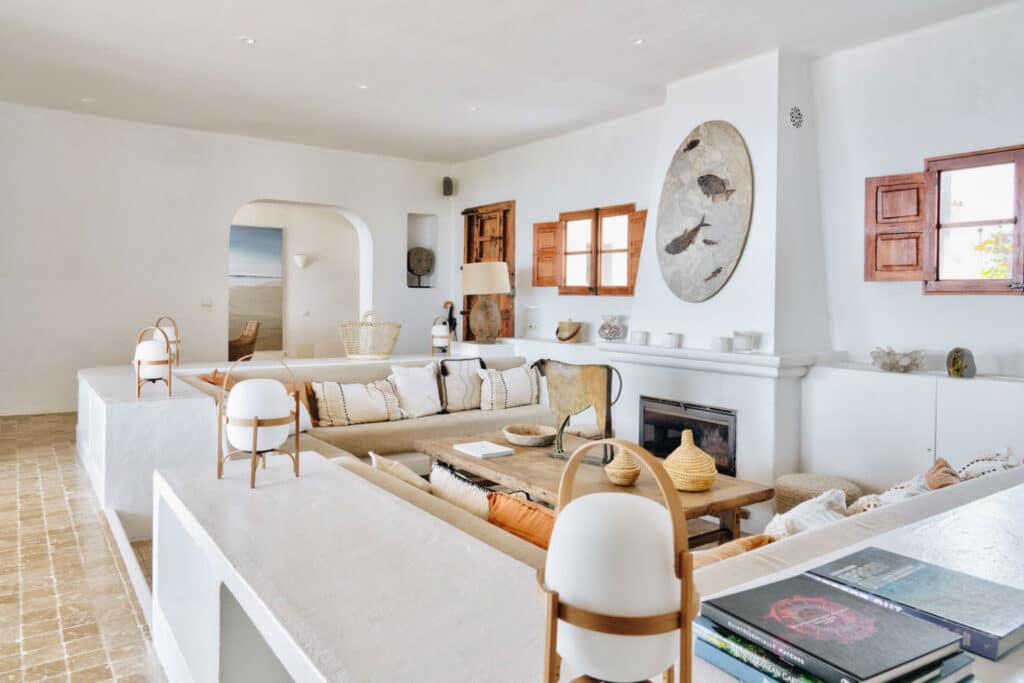 Spacious living room with white walls and earthy tones reminiscent of luxury travel. Features a large L-shaped sofa, wooden coffee table with decor, wicker baskets, wall art, and a fireplace. Natural light enters through small windows with wooden shutters. David Pearson Travel