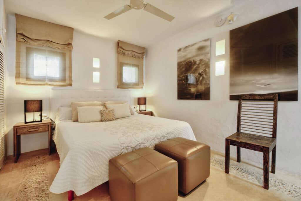A cozy bedroom reminiscent of luxury travel, with a large bed adorned in decorative pillows and a white bedspread. Two brown ottomans sit at the foot of the bed, surrounded by wooden shutters, a rustic chair, two lamps, and framed pictures on the walls. David Pearson Travel