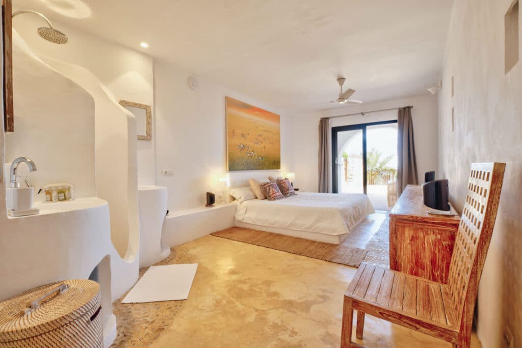 A bright, minimalist bedroom designed for luxury travel features a large bed with colorful pillows, a painting above, an open shower area, a rustic wooden chair and table with a TV, and a glass door leading to a balcony overlooking swaying palm trees. David Pearson Travel