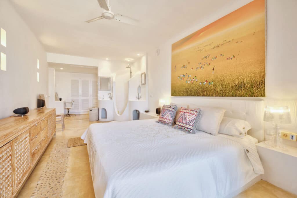 A bright bedroom with beige walls and flooring exudes a luxury travel aura. The bed boasts white linens and patterned pillows, while a large artwork of a field scene hangs above. A wooden dresser, ceiling fan, and modern wall sconces complement the space, with natural light pouring through the window. David Pearson Travel