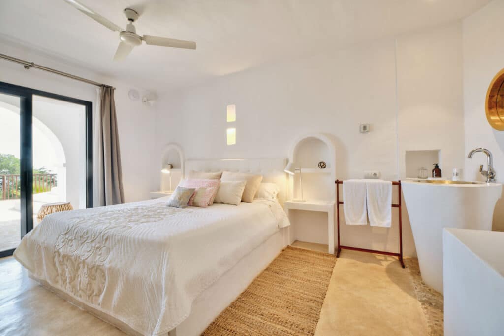 A bright, airy bedroom perfect for luxury travel enthusiasts, featuring a large bed adorned with white linens and decorative pillows. The room boasts a ceiling fan, arched windows, a small sink, towel rack, and wicker chair. Natural light elegantly fills the space through glass doors. David Pearson Travel