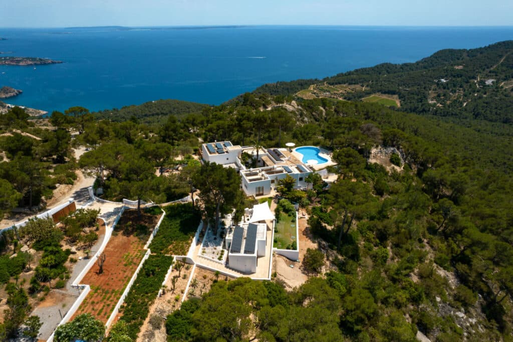 Aerial view of a luxury travel villa nestled amidst lush greenery on a hillside, overlooking the ocean. The property boasts a pool with a deck and solar panels on the roof, with pathways winding through gardens and trees. The sea stretches to the horizon under a clear sky. David Pearson Travel