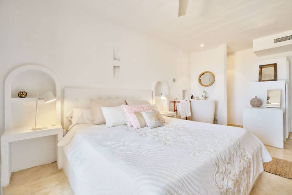 Bright, minimalist bedroom with white walls, a large bed covered in a white quilt, and pink accent pillows reminiscent of luxury travel accommodations. Two bedside tables with lamps flank the bed. A round mirror and decorative vases add subtle decor to the serene space. David Pearson Travel