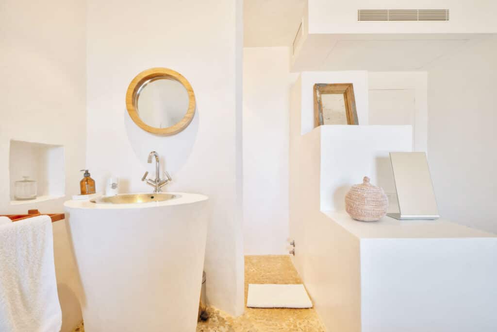 A touch of luxury travel-inspired elegance, this minimalist white bathroom features a round mirror above a sleek, modern basin. The wooden-topped counter holds a wicker basket and towel rack, while the natural stone floor enhances the serene atmosphere. David Pearson Travel