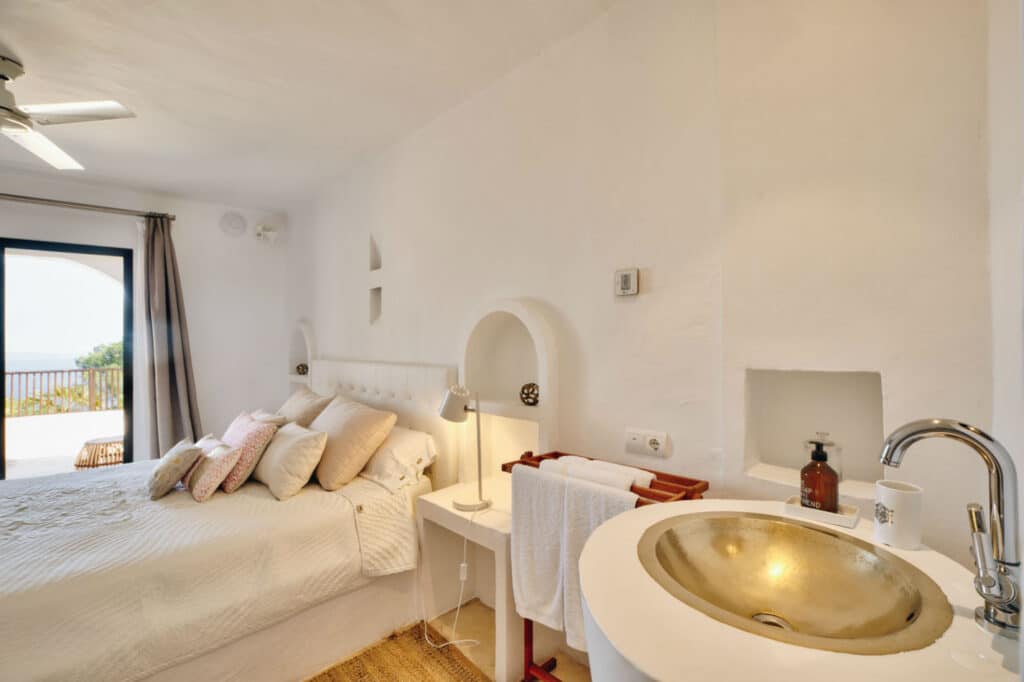 Bright bedroom with white walls, a plush bed adorned with multiple pillows, and a small round sink adjacent to it. A window offers a view of the balcony and lush greenery. A bedside table holds a lamp, while a towel rack stands nearby—perfect for those appreciating luxury travel comforts. David Pearson Travel