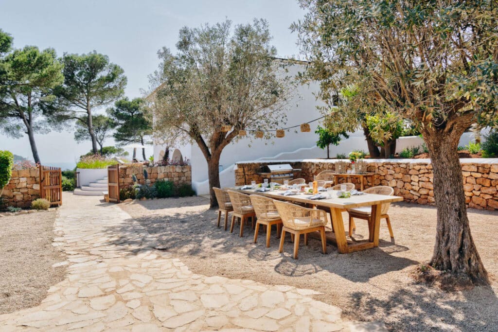 The outdoor dining area exudes luxury travel vibes, featuring a wooden table and wicker chairs set for a meal under large trees. Enclosed by stone walls and lush greenery, a stone pathway beckons toward a wooden gate. A pristine white building stands elegantly against the clear sky. David Pearson Travel