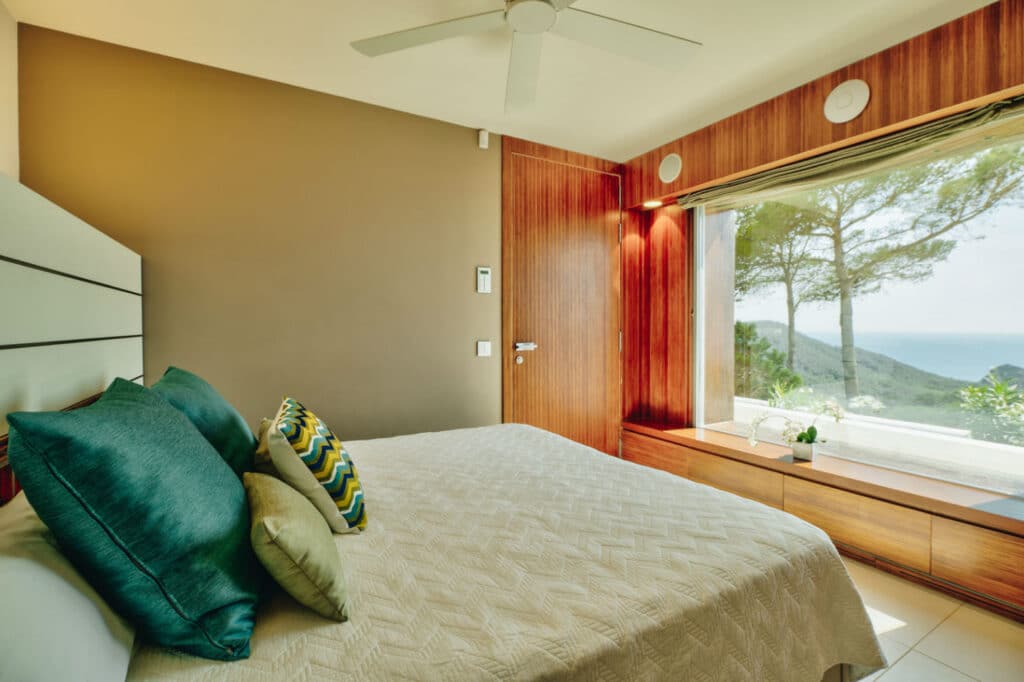 A modern bedroom, ideal for luxury travel enthusiasts, boasts a large window framing a scenic view of trees and the ocean. It features a neatly made bed with green and patterned pillows, a ceiling fan, and wooden accents on the walls and window seat. David Pearson Travel