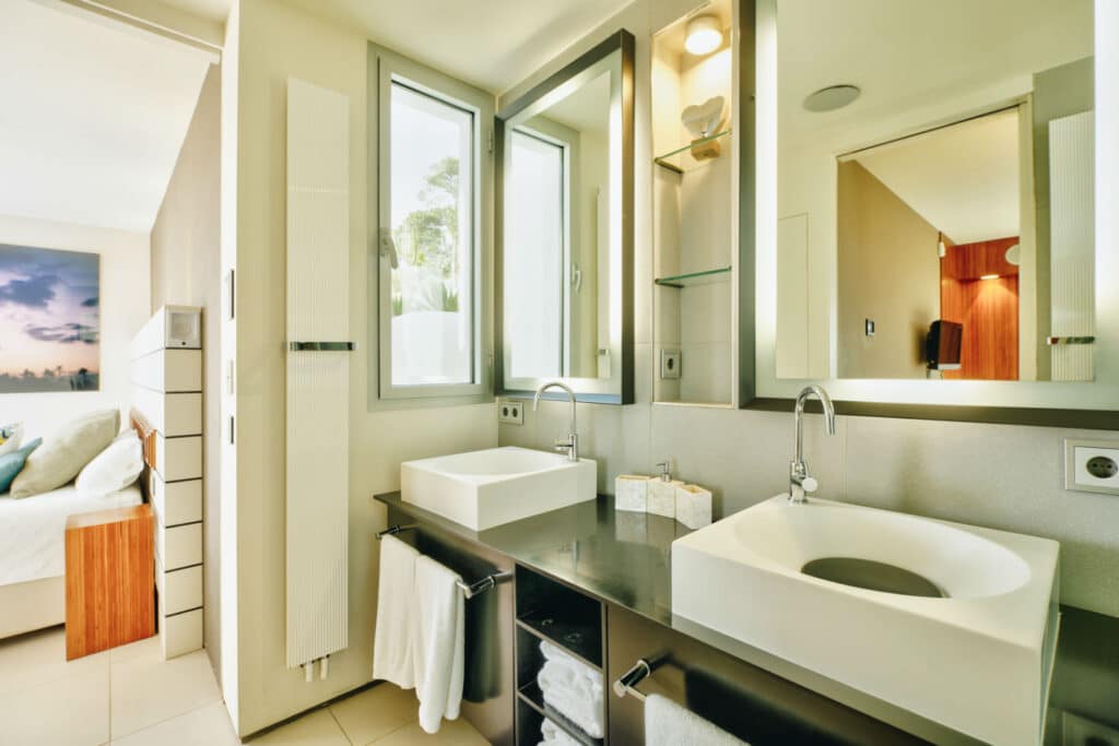 Modern bathroom with two square sinks on a black countertop, large mirrors, and shelves. A window on the left allows natural light. Towels are neatly placed below the sinks. The space evokes luxury travel vibes, with a bedroom featuring wooden accents visible through an open doorway. David Pearson Travel