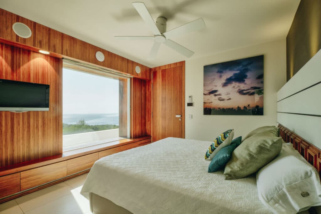This modern bedroom, perfect for luxury travel enthusiasts, features a wooden accent wall and a large window showcasing an ocean view. A flat-screen TV faces the white bed adorned with decorative pillows under a ceiling fan, complementing the framed artwork beside the window. David Pearson Travel