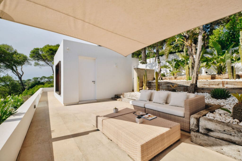 Luxury Travel: Experience an outdoor patio with a beige woven couch and coffee table under a large canopy, embraced by white walls and lush greenery. A potted plant and decorative rocks add natural elements, while a door to the building hints at opulent journeys beyond. David Pearson Travel