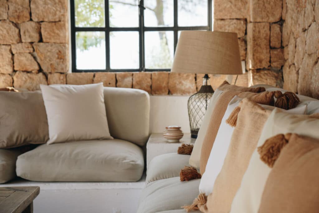 Cozy living room with a beige sofa adorned with tasseled pillows, set against a stone wall. A large lamp and a stack of bowls sit on a table by the window, where natural light pours in, evoking the elegance of luxury travel in your own home. David Pearson Travel