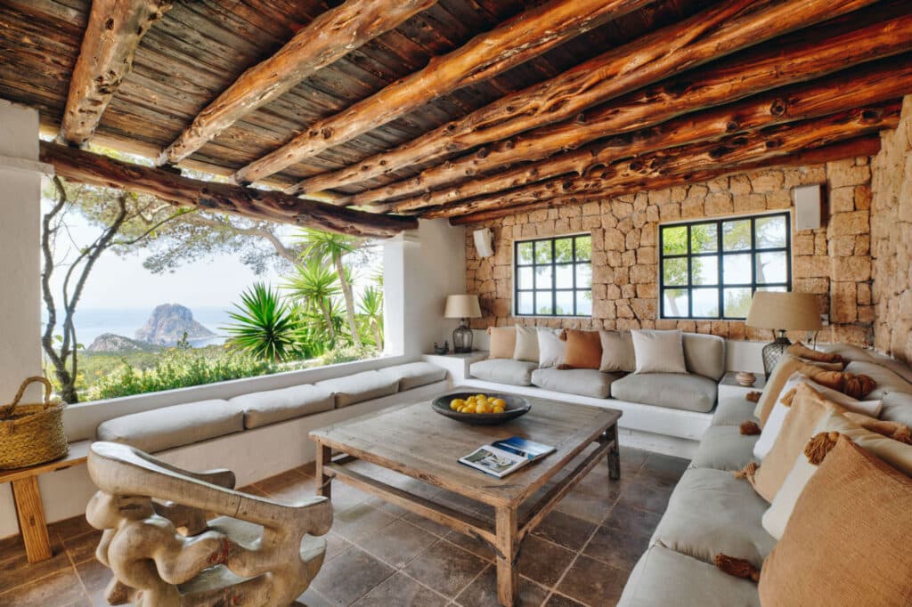 Rustic living room with wooden ceiling beams, stone walls, and large windows overlooking lush greenery and a distant sea view—a perfect setting for luxury travel. Two white sofas are positioned around a wooden coffee table with a bowl of lemons. A carved wooden chair and basket complete the decor. David Pearson Travel