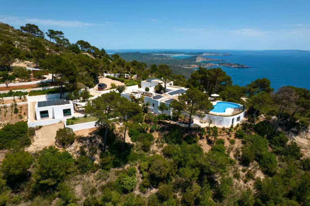 Aerial view of a white villa surrounded by lush greenery on a hillside, featuring a circular pool—a true luxury travel escape. The villa overlooks a vast blue sea and distant islands under a clear blue sky. David Pearson Travel