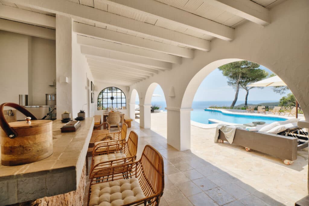 Experience luxury travel with a Mediterranean-style patio featuring wicker chairs at a stone bar counter. Overlooking an infinity pool and ocean view, arched openings let sunlight in, creating a bright and inviting atmosphere, while poolside loungers rest under a tree. David Pearson Travel