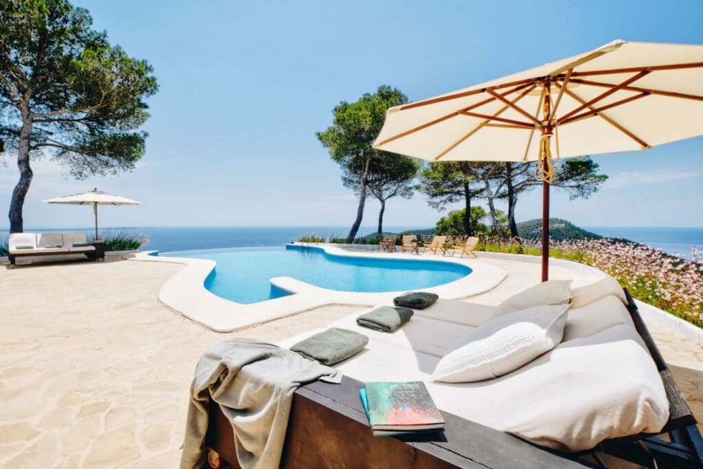 A luxurious poolside scene perfect for luxury travel enthusiasts overlooks the sea, with a white lounge chair, umbrellas, and trees framing the view. Towels and a book are placed on the chair. The blue sky and ocean create a serene atmosphere. David Pearson Travel