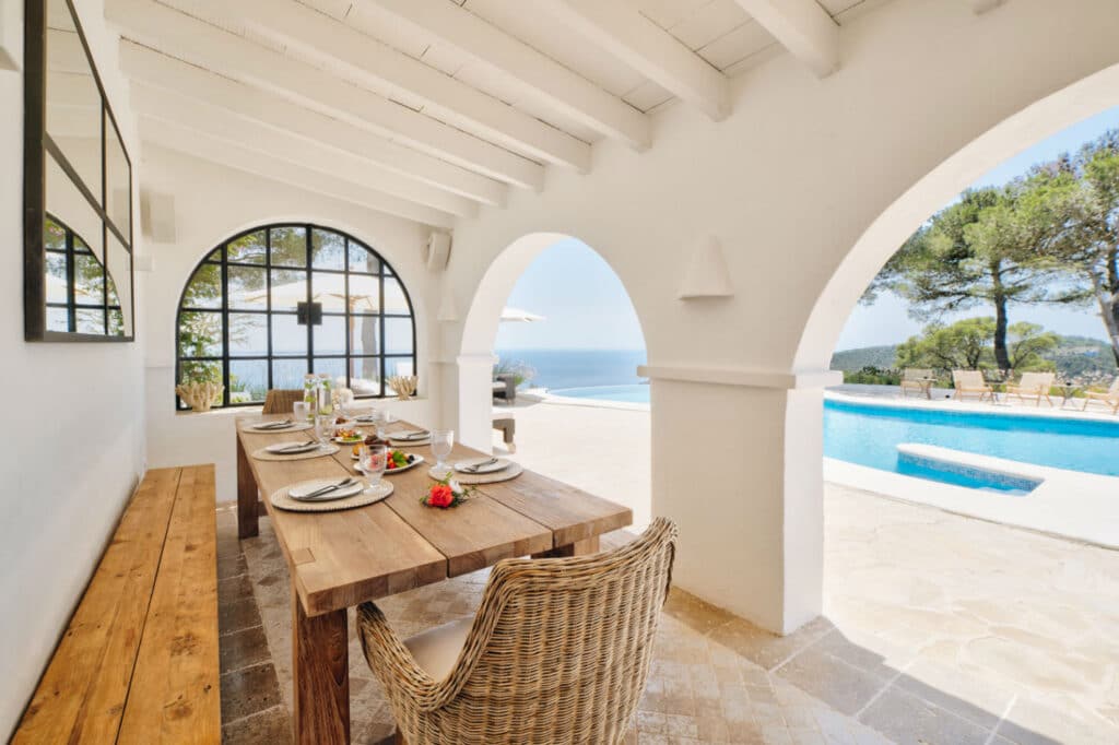 A serene outdoor dining area features a wooden table set for a meal beneath a white pergola. Arched openings reveal a stunning view of a pool, lush greenery, and the ocean, embodying the essence of luxury travel with its tranquil and inviting atmosphere. David Pearson Travel