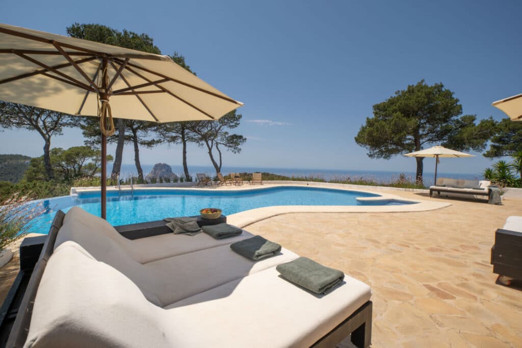 Experience the epitome of luxury travel at this poolside patio, complete with sun loungers and large umbrellas under a clear sky. Overlooking the ocean and surrounded by trees, towels are neatly placed for your comfort, enhancing the tranquil atmosphere perfect for ultimate relaxation. David Pearson Travel