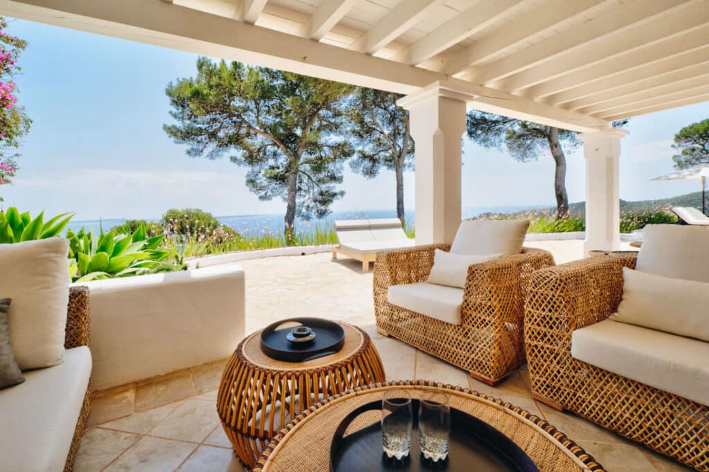 Discover a slice of luxury travel right at home with an outdoor patio featuring wicker furniture, including a sofa and chairs, comfortably nestled under a white pergola. The scene, with its round table holding a tray of glasses, overlooks lush greenery and trees beneath the blue sky. David Pearson Travel